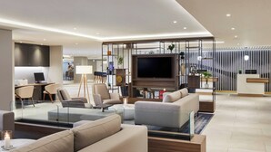 A modern living room with a flat-screen TV, comfortable seating, a glass coffee table, and a kitchen area in the background. - Courtyard by Marriott London Heathrow Airport (Hayes)