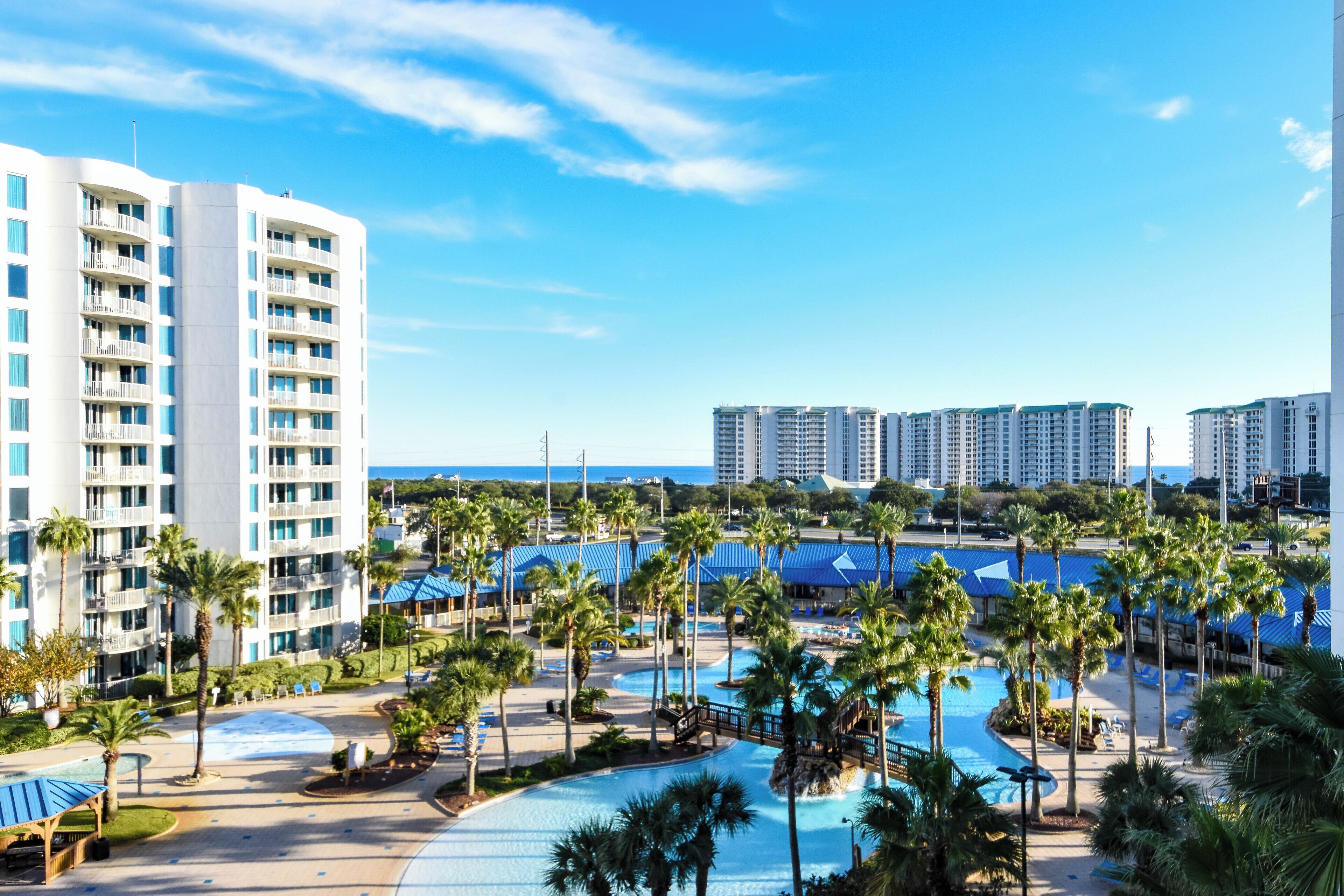 A resort with multiple high-rise buildings, a central pool area surrounded by palm trees, and a clear view of the ocean.