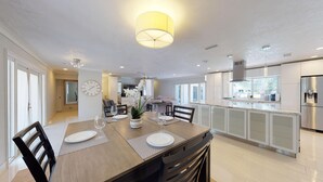Een moderne keuken met een centraal kookeiland, een eethoek met een tafel gedekt voor twee personen, en een zithoek met een bank en een televisie.