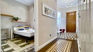Quarto de hotel com cama, escrivaninha, cadeira, planta em vaso, quadro e piso estampado.
