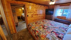 A cozy log cabin bedroom with a quilted bedspread, a television, and a window with curtains. - Cabin within sight distance of Lake Superior, Autrain River. Free Kayak rental (Au Train)