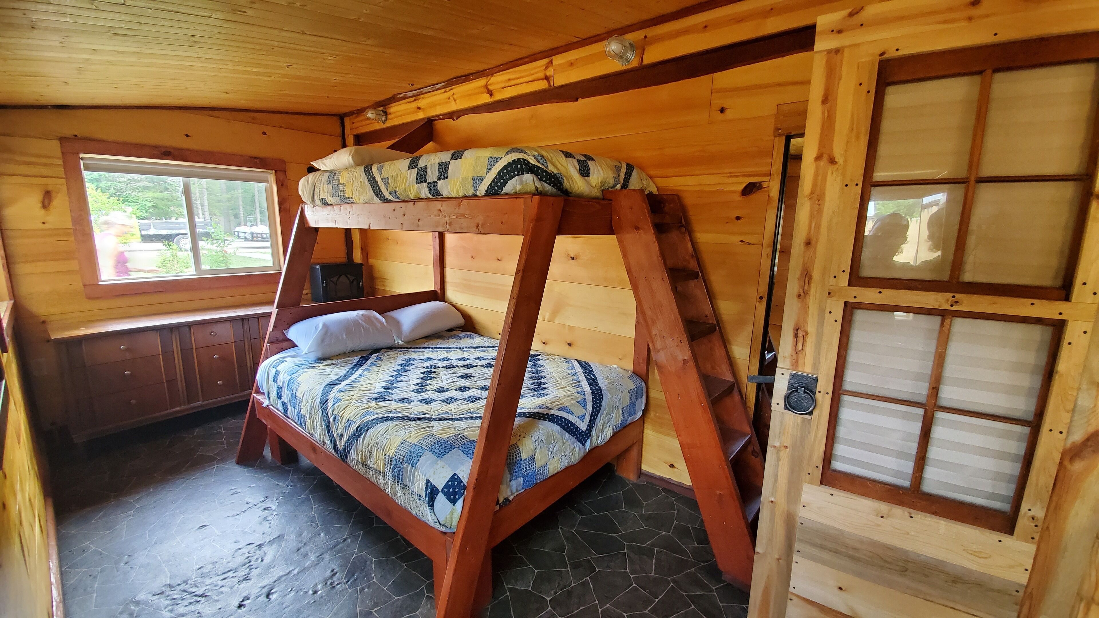 A bunk bed with a patterned bedspread, wooden walls, a window with a view, and a door with a window.