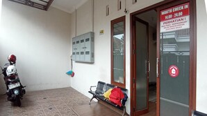 A waiting area with a bench, a motorcycle, and a door with a sign indicating check-in hours. - OYO 3314 Syariah House (Purwokerto)