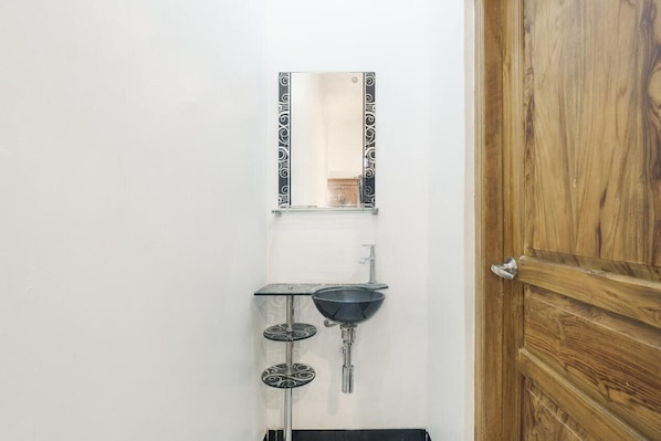 Ein Badezimmer mit Holztür, einem wandmontierten Spiegel und einem Waschbecken mit schwarzem Becken sowie zwei runden, verzierten Ablageflächen darunter.