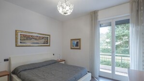 A bedroom with a bed, a nightstand, a chandelier, a window with curtains, and two framed pictures on the wall. - Aura Home Sirmione - 2 bedrooms apartment (Sirmione)