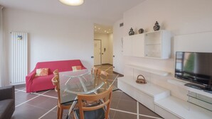 A modern living room with a glass dining table, a red sofa, a white cabinet, and a flat-screen TV. - Aura Home Sirmione - 2 bedrooms apartment (Sirmione)