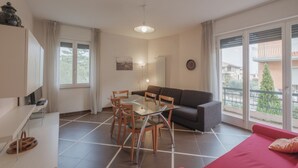 A modern living room with a glass dining table, wooden chairs, a grey sofa, and a red sofa. There is a balcony with a view of buildings and greenery. - Aura Home Sirmione - 2 bedrooms apartment (Sirmione)
