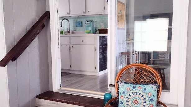 Una escalera conduce a una cocina con armarios blancos, una silla de madera con cojín estampado y un cartel que dice 'Welcome'.