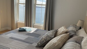 Un dormitorio con una cama grande, varios almohadones, vistas al agua a través de amplios ventanales y una lámpara en la mesita de noche.