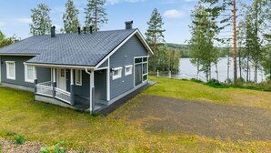 Una casa de dos pisos con fachada gris y detalles blancos, un porche cubierto y vista a un lago rodeado de árboles.
