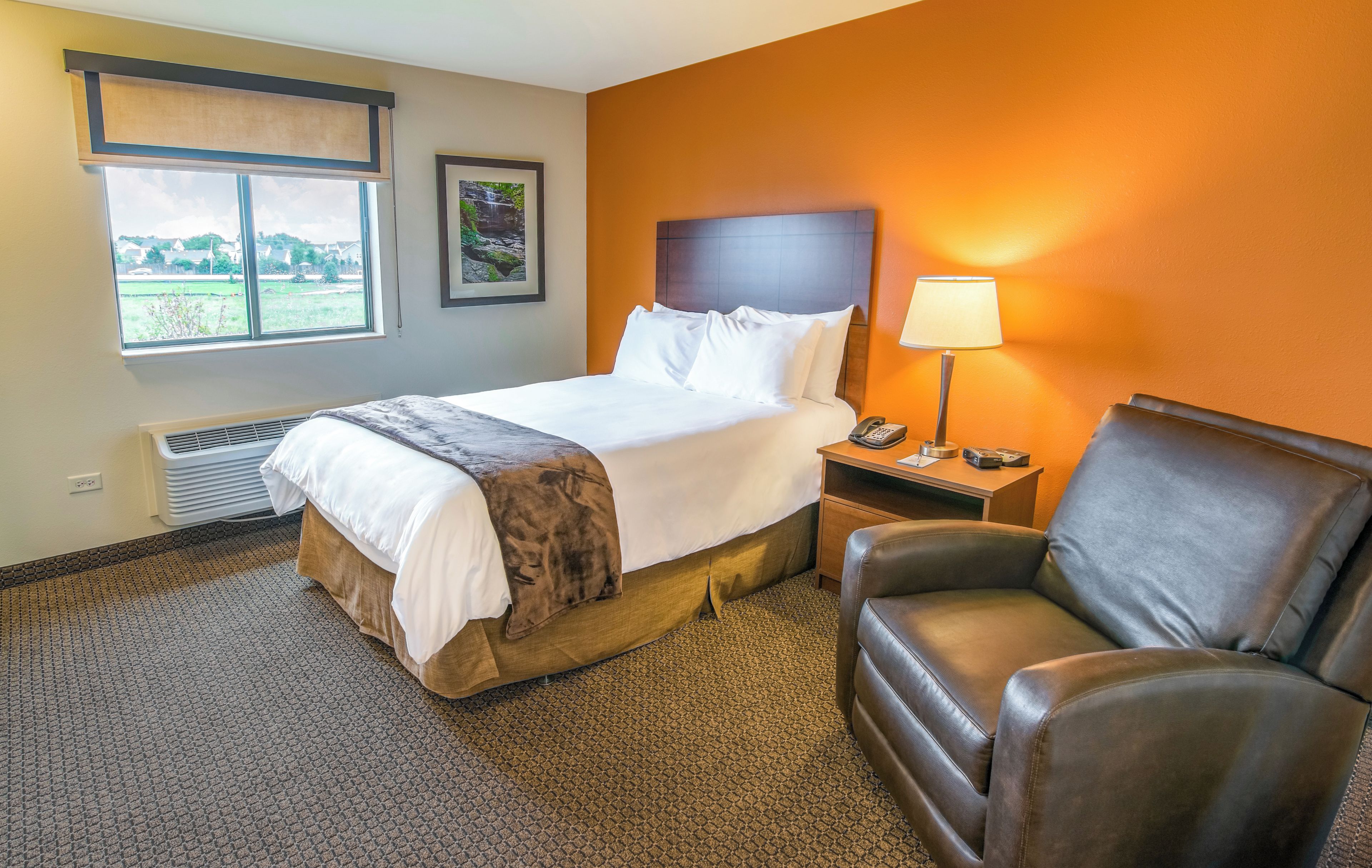 A hotel room with a bed, a chair, a nightstand with a lamp, and a window with a view of greenery.