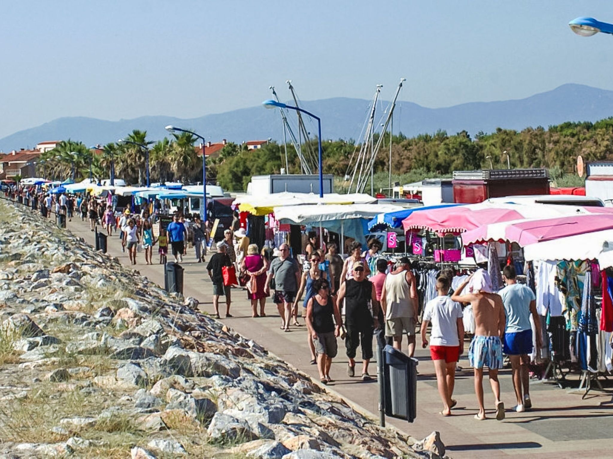 Ein belebter Markt im Freien mit Menschen, die auf einem gepflasterten Weg entlanggehen, Händler mit bunten Sonnenschirmen und einer felsigen Küstenlinie im Hintergrund.