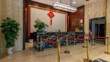 A hotel reception area with a marble counter, red ropes, and potted plants.