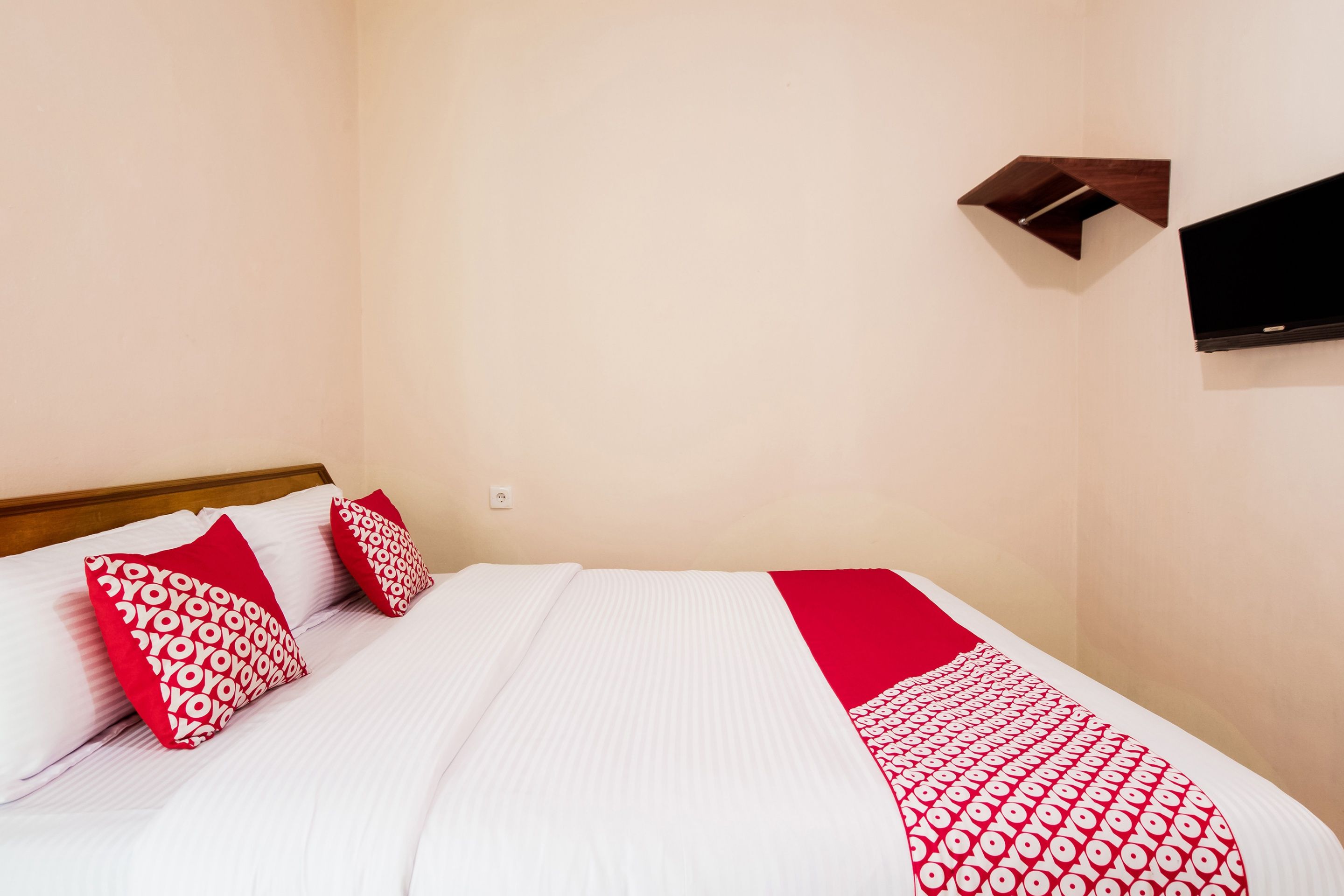 A neatly made bed with white and red bedding, a mounted TV on the wall, and a power outlet on the wall.