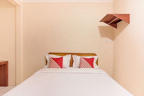 A neatly made bed with white bedding and two red patterned pillows, a wooden headboard, and a small wooden shelf on the wall.