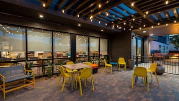 An outdoor patio area with yellow chairs and tables, large windows, and a brick floor.