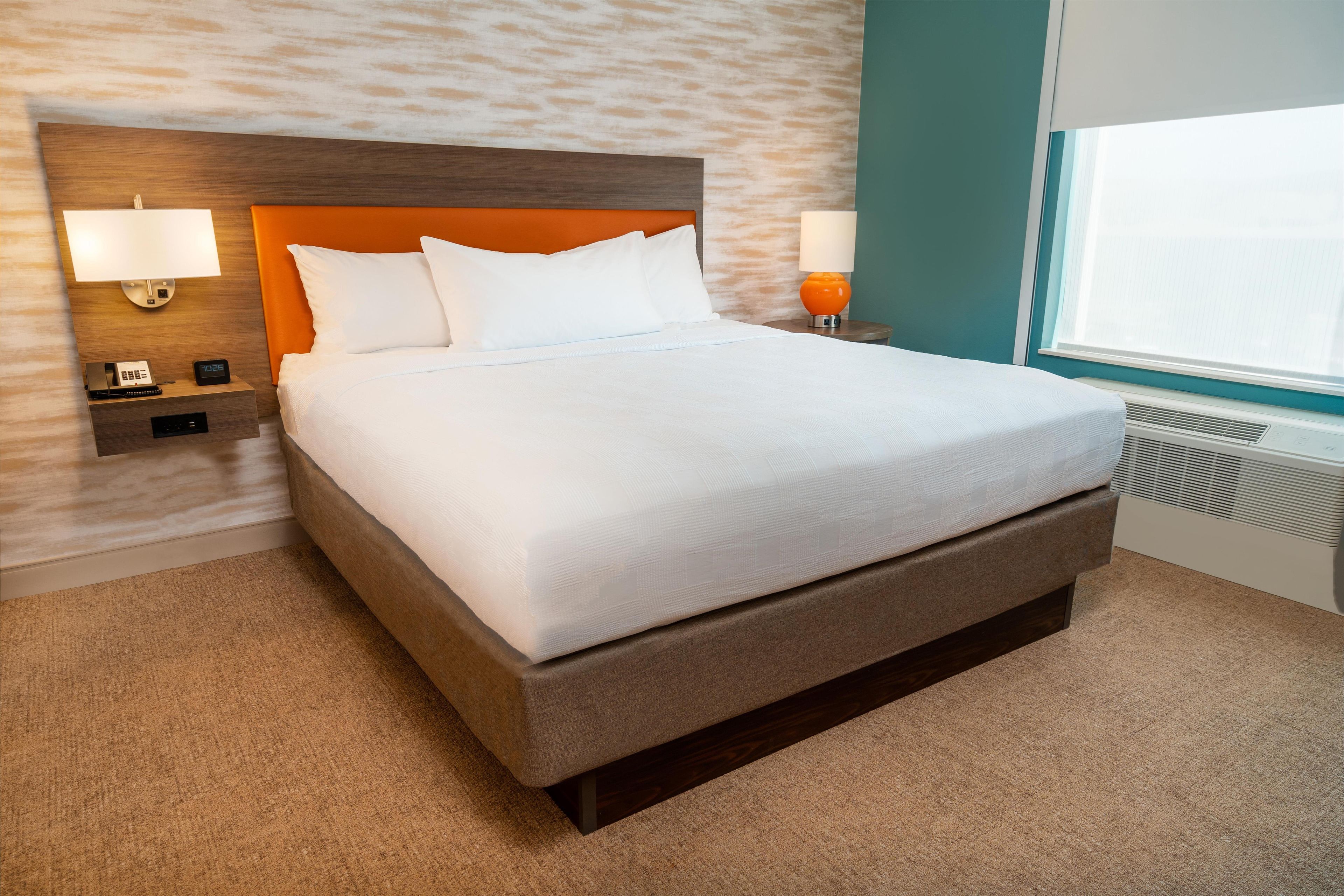 A neatly made bed with white linens in a hotel room with a textured wall, a wooden headboard, and a window with blinds.