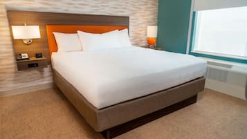 A neatly made bed with white linens in a hotel room with a textured wall, a wooden headboard, and a window with blinds.