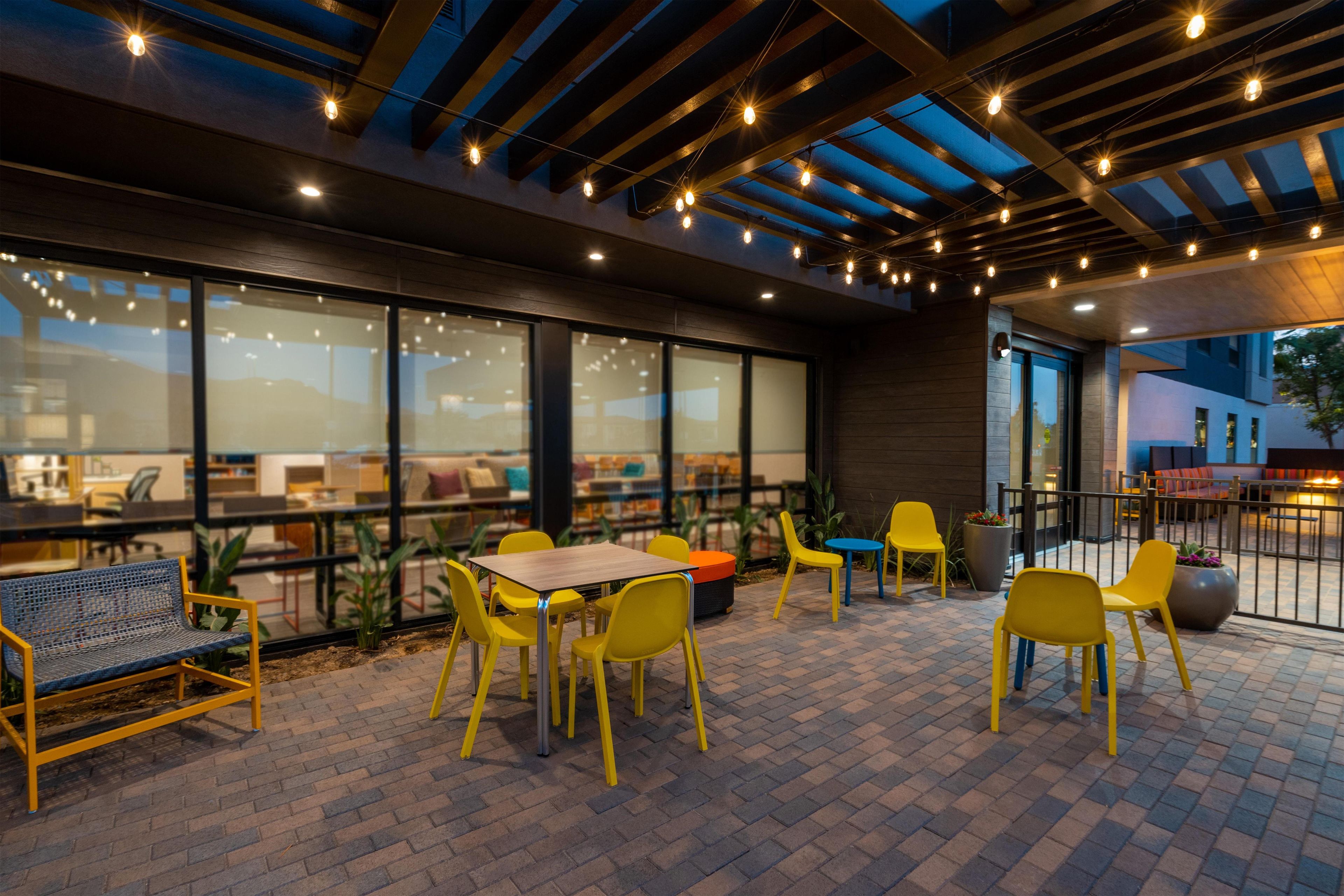 An outdoor seating area with yellow chairs and tables, large windows, and a covered section with string lights.