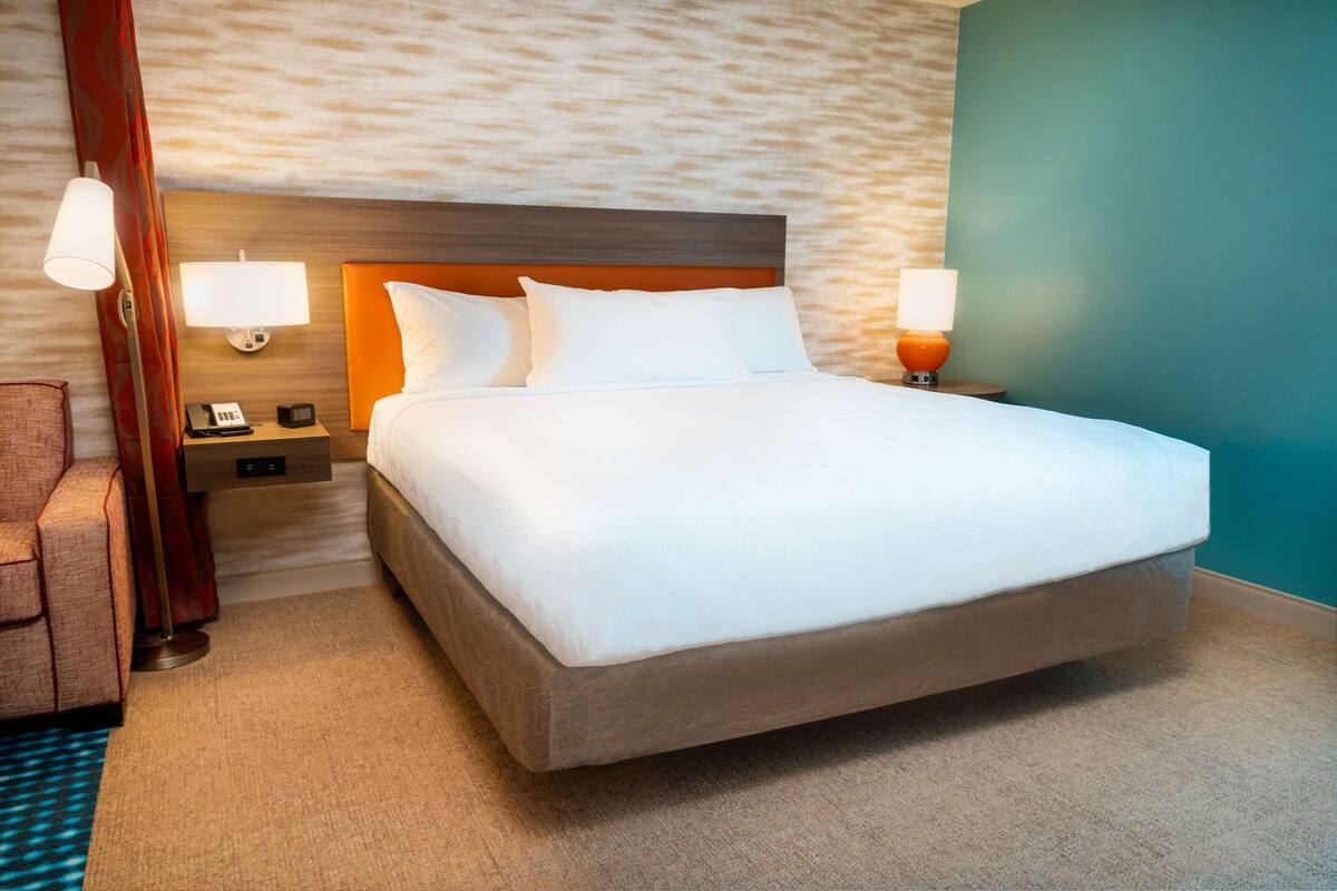 A large bed with white bedding, a wooden headboard, two bedside lamps, a nightstand with a telephone, and a red chair.