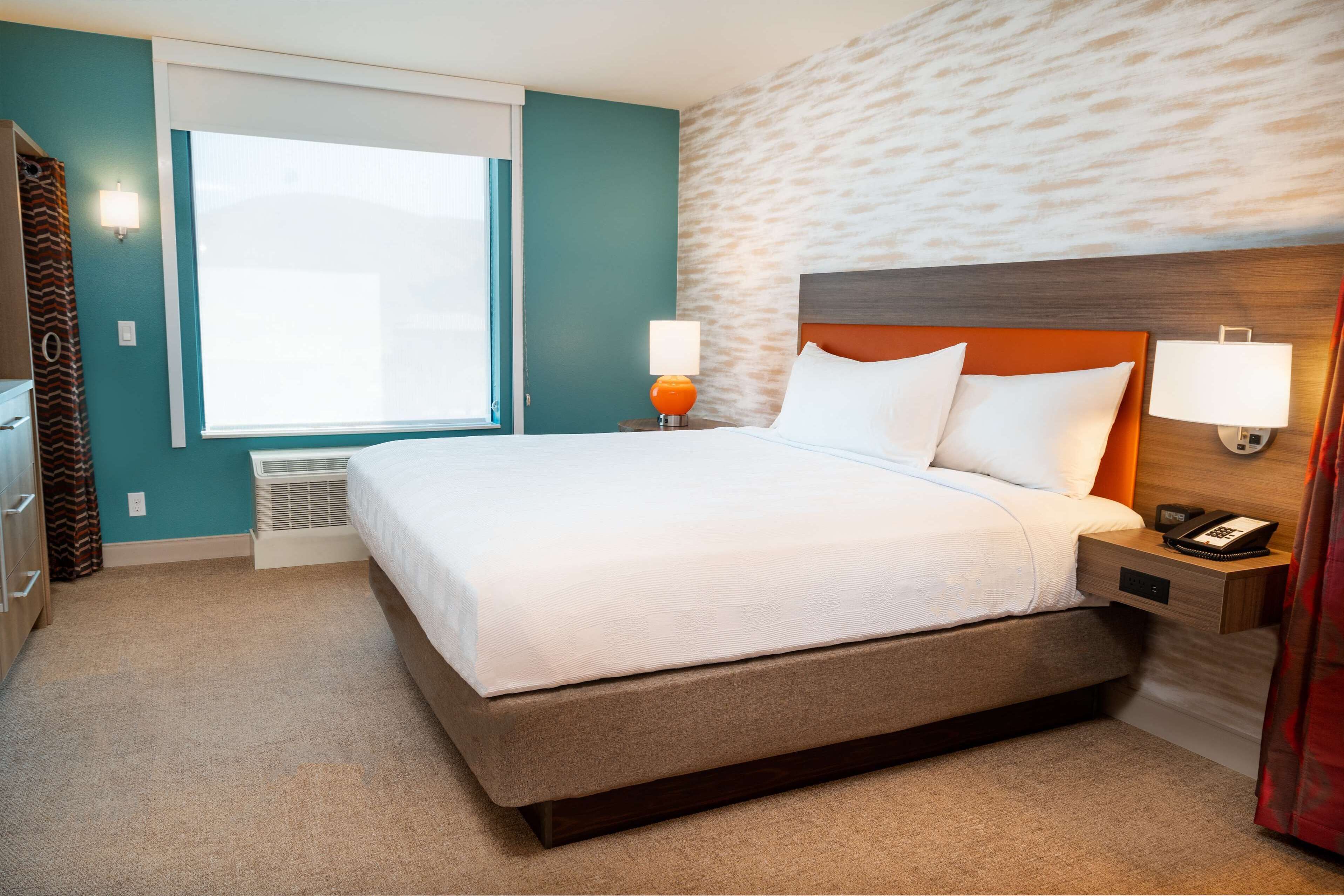Une chambre d’hôtel avec un grand lit, une fenêtre avec des rideaux, une lampe fixée au mur et un thermostat.