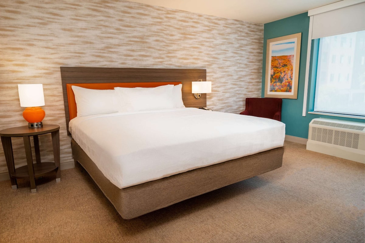 A hotel room with a large bed, two bedside lamps, a red chair, a window with a view, and a wall-mounted air conditioner.