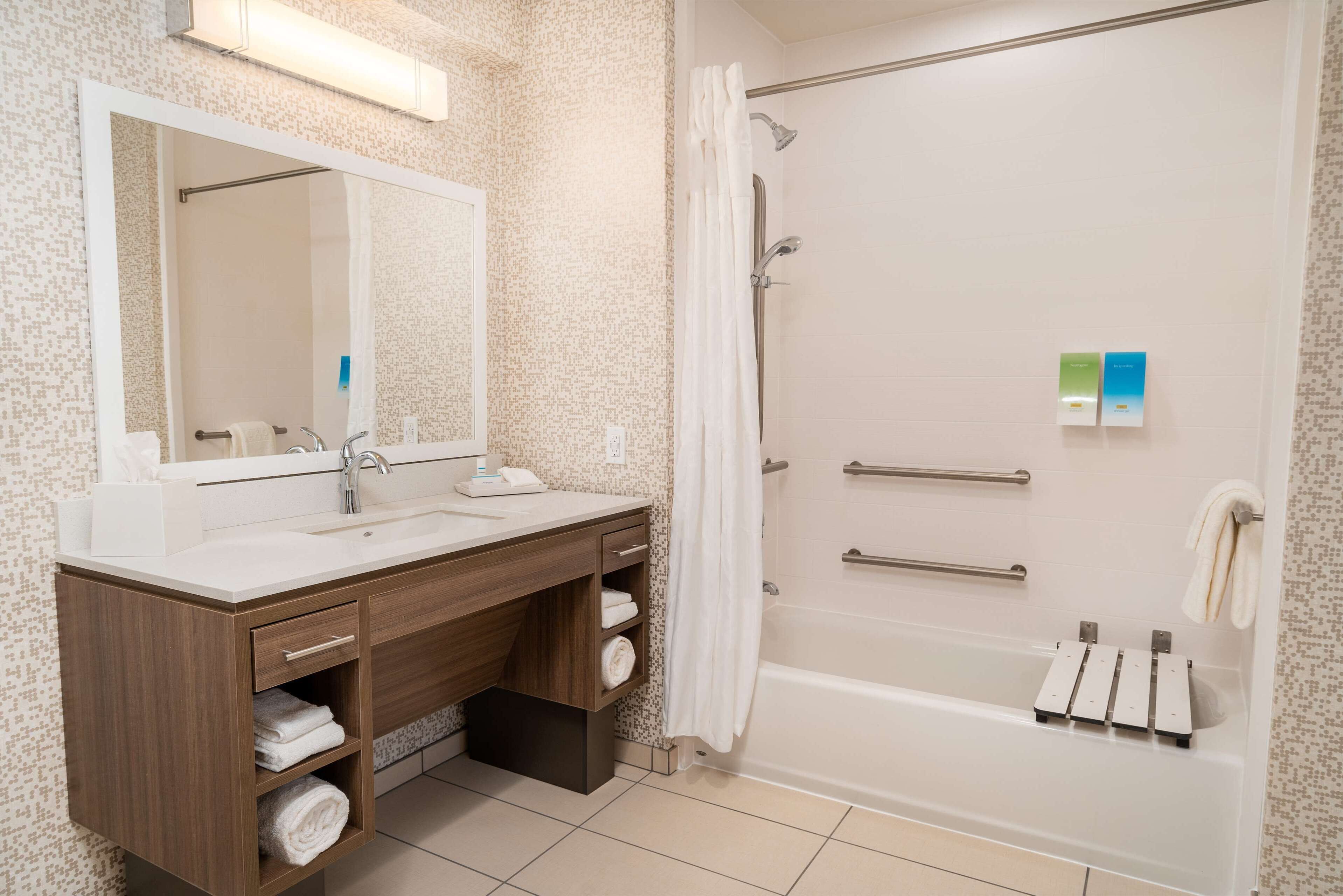Une salle de bain moderne avec un grand miroir, deux vasques, une douche équipée de barres d’appui et des serviettes soigneusement rangées.