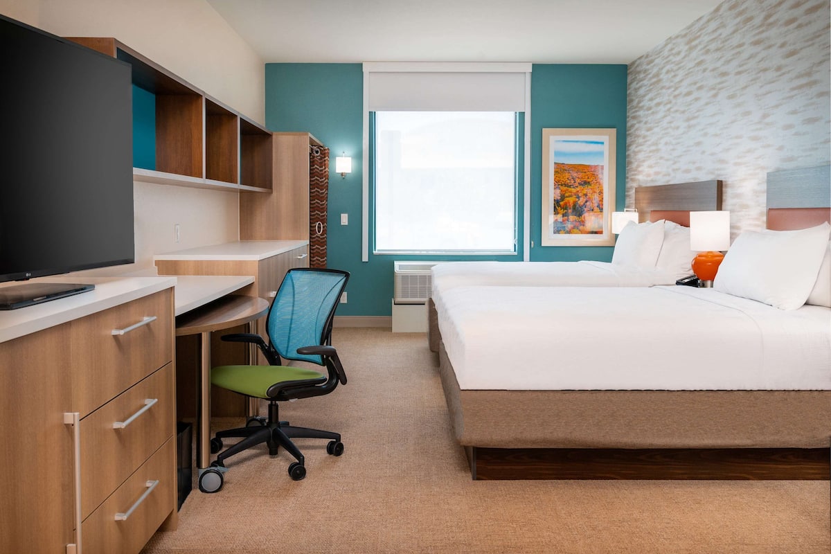 A hotel room with a bed, desk, chair, TV, and artwork on the wall.