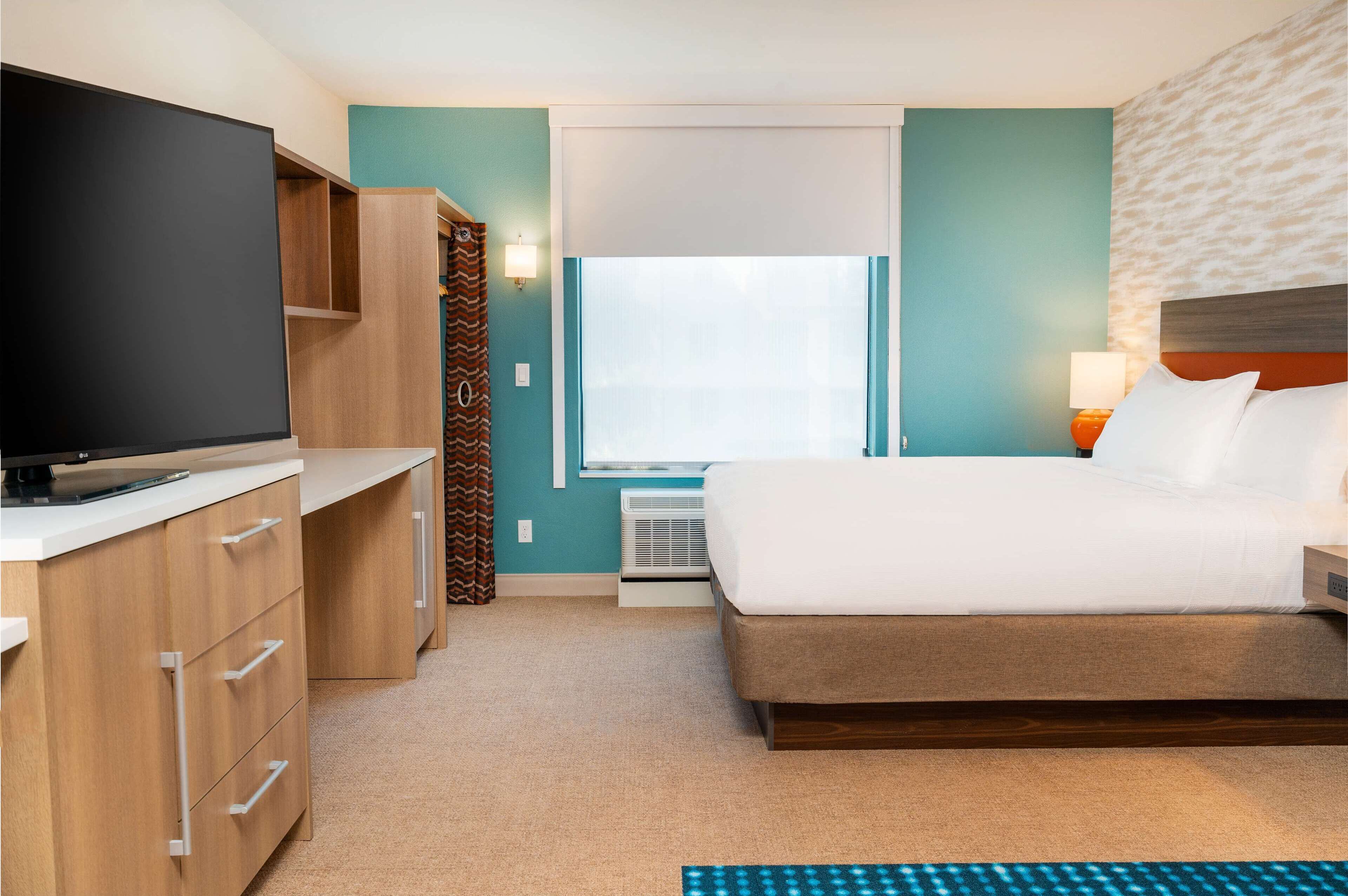 Una habitación de hotel con cama, televisor, escritorio con cajones, ventana con cortinas y un calentador en la pared.
