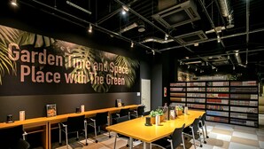 A conference room with a large banner reading "Garden Time and Space Place with The Green," a bookshelf labeled "Magazine Corner," and a checkered floor pattern. - Design Hotel Blax (Hachioji)