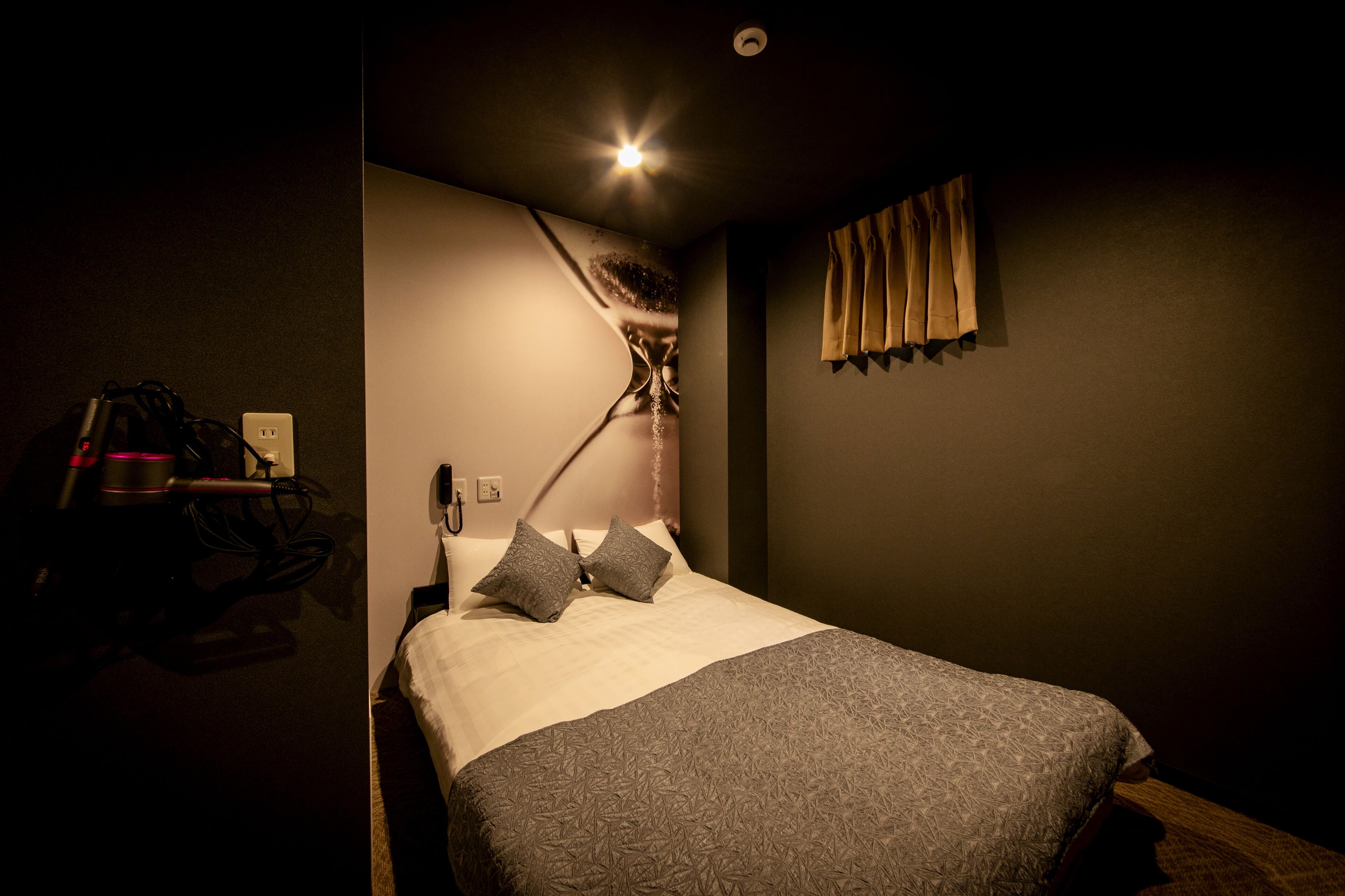 A dimly lit hotel room with a bed, a desk with a lamp, and a curtain on the window.