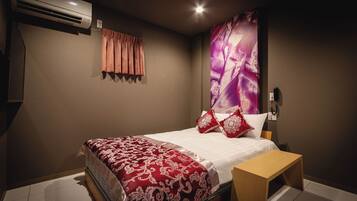 A bedroom with a bed, a bedside table, an air conditioning unit, a wall-mounted light, and a large floral artwork.