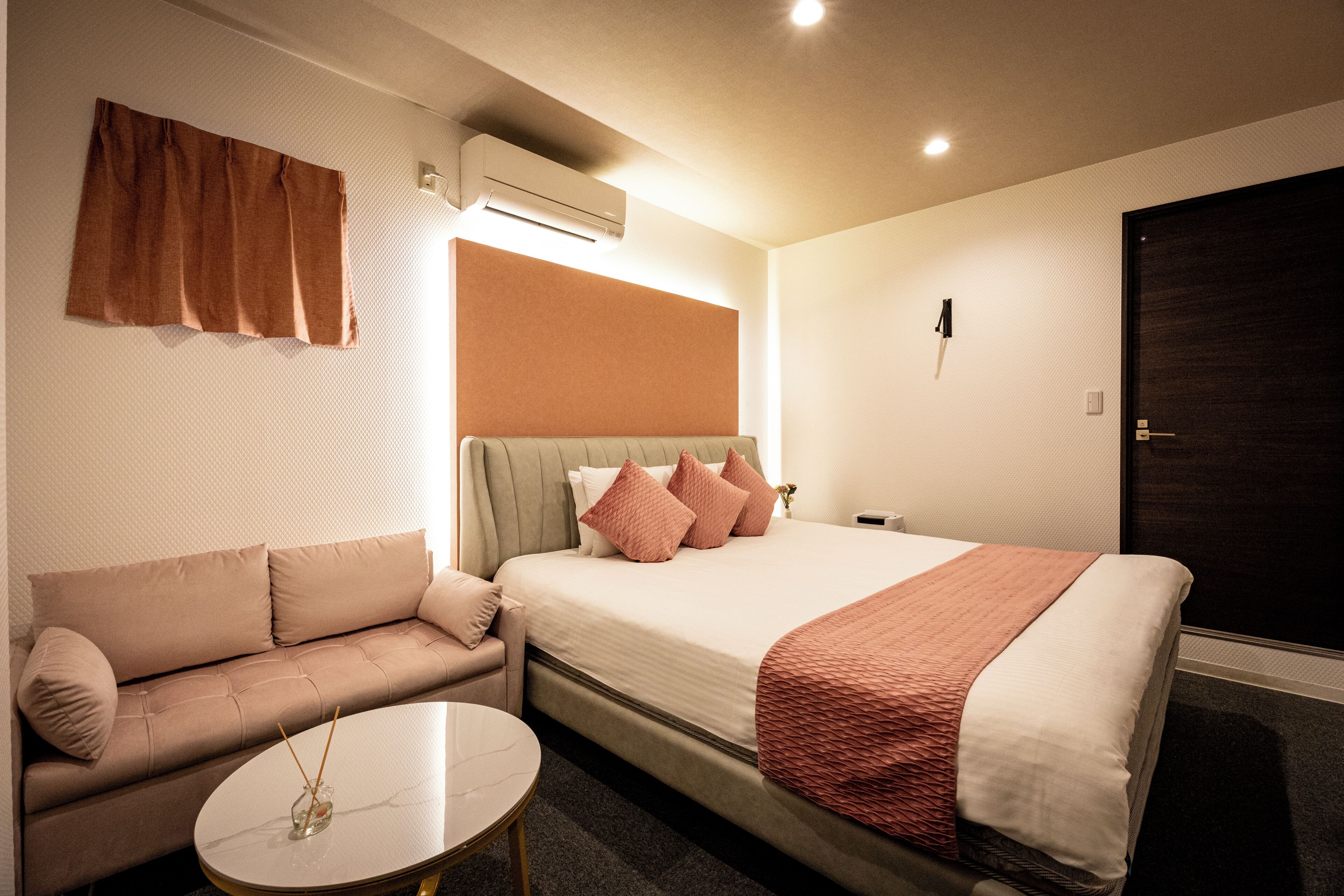 A hotel room with a bed, a sofa, a round table, and an air conditioning unit.