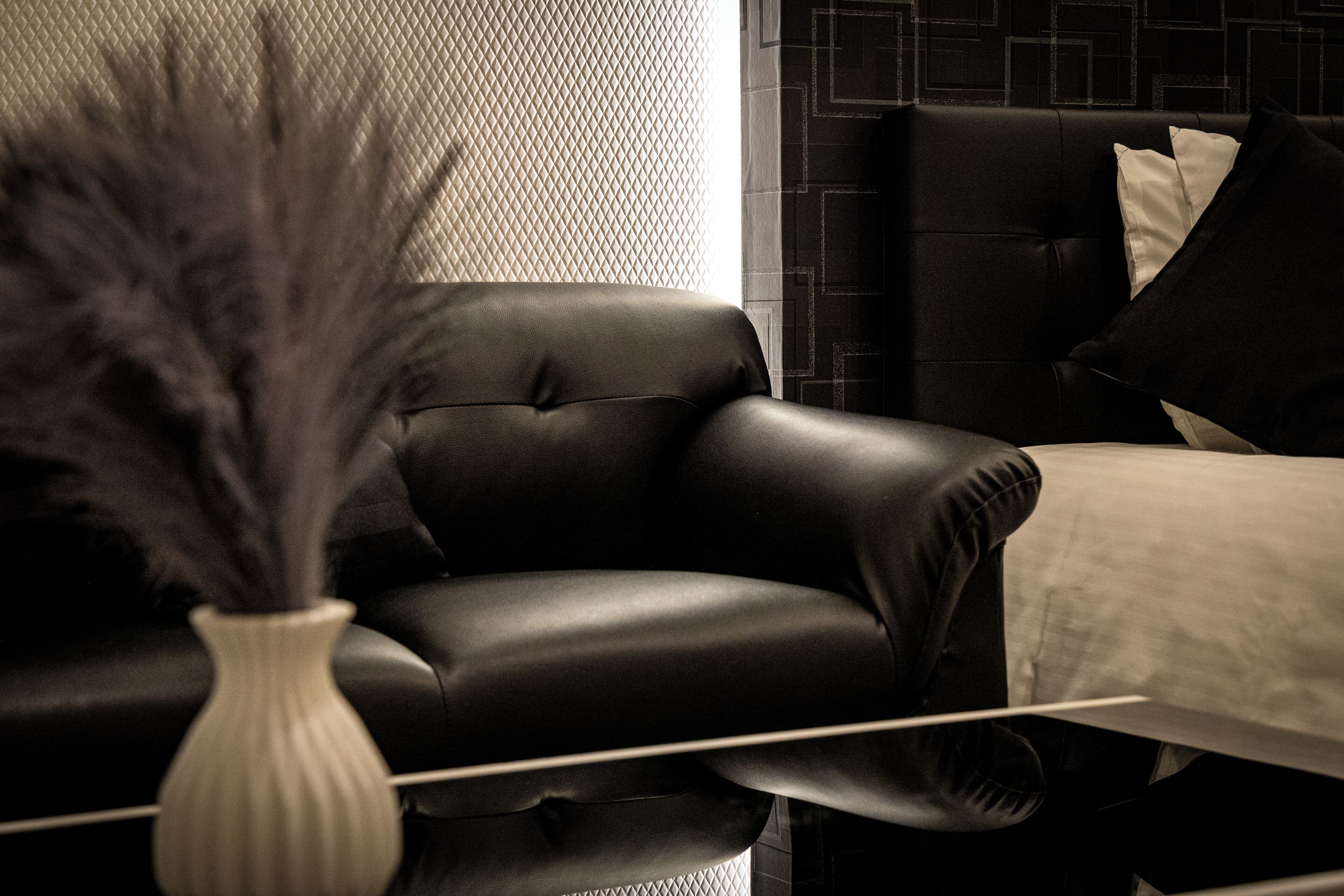 Um sofá de couro preto, um vaso branco com detalhes decorativos em preto e uma parede estampada.