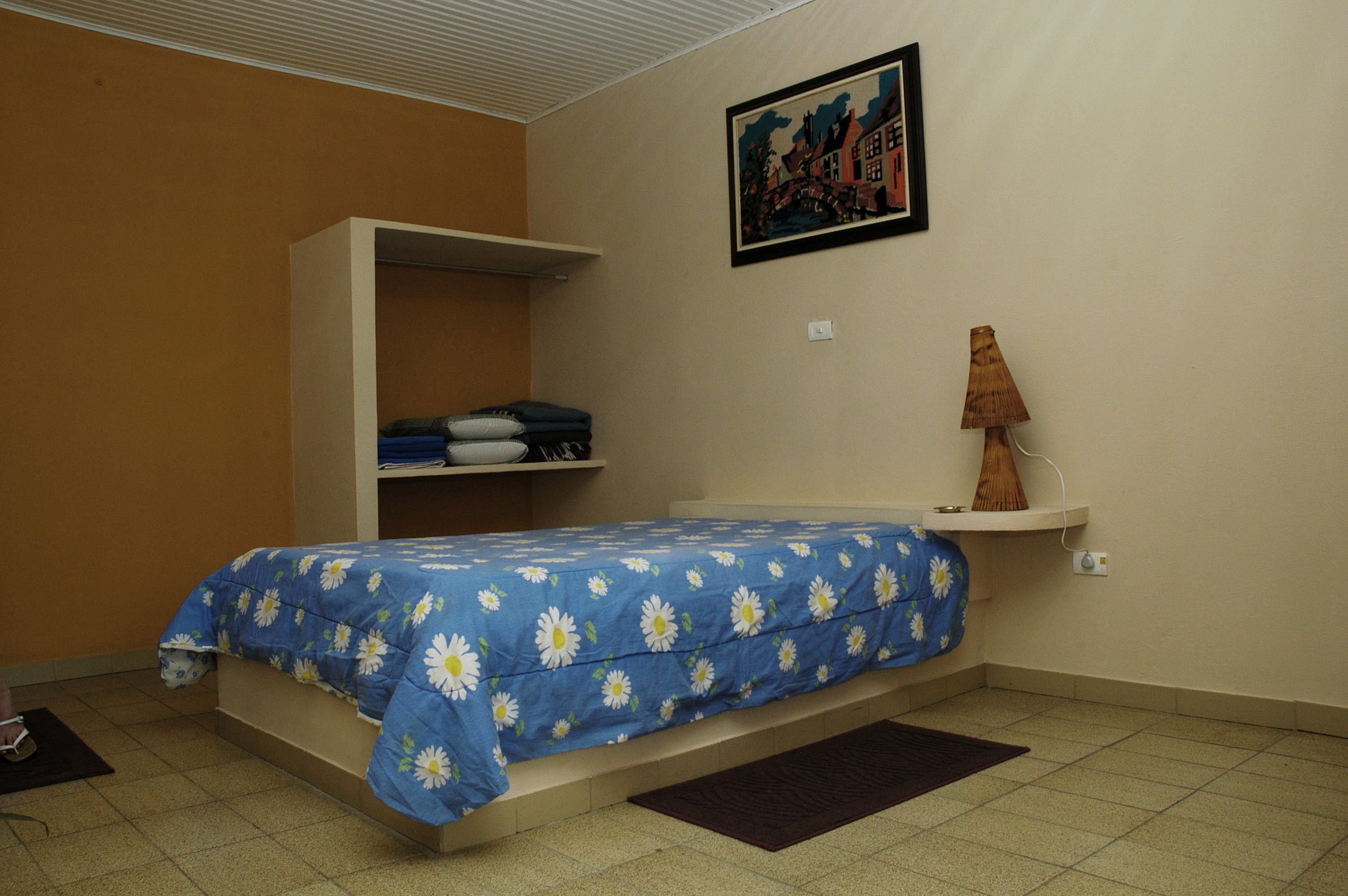 A bedroom with a bed, a wardrobe, a shelf with folded clothes, a lamp, and a painting on the wall.
