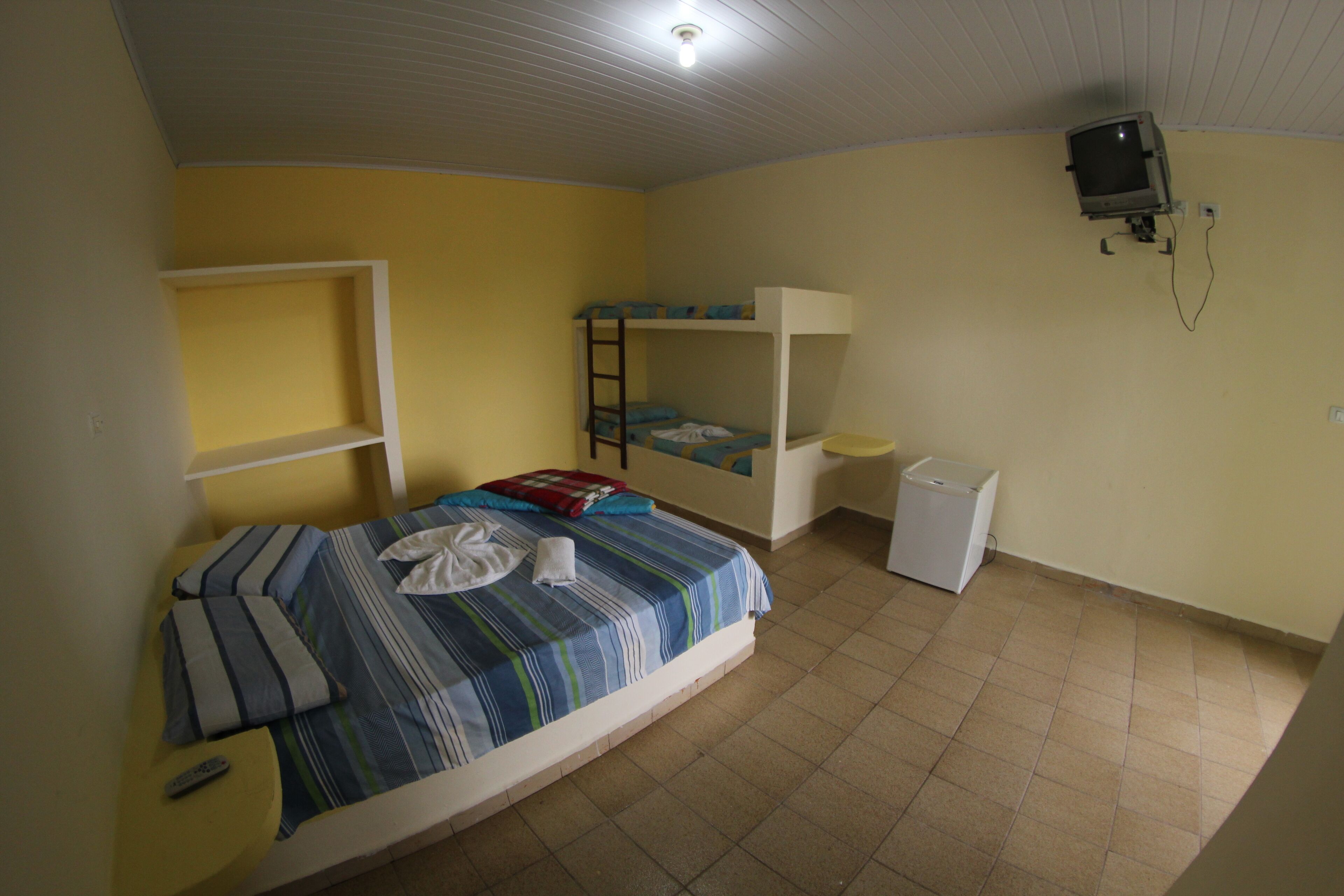 Quarto estilo dormitório com beliches, uma pequena escrivaninha, uma cadeira, TV fixada na parede e um refrigerador branco.