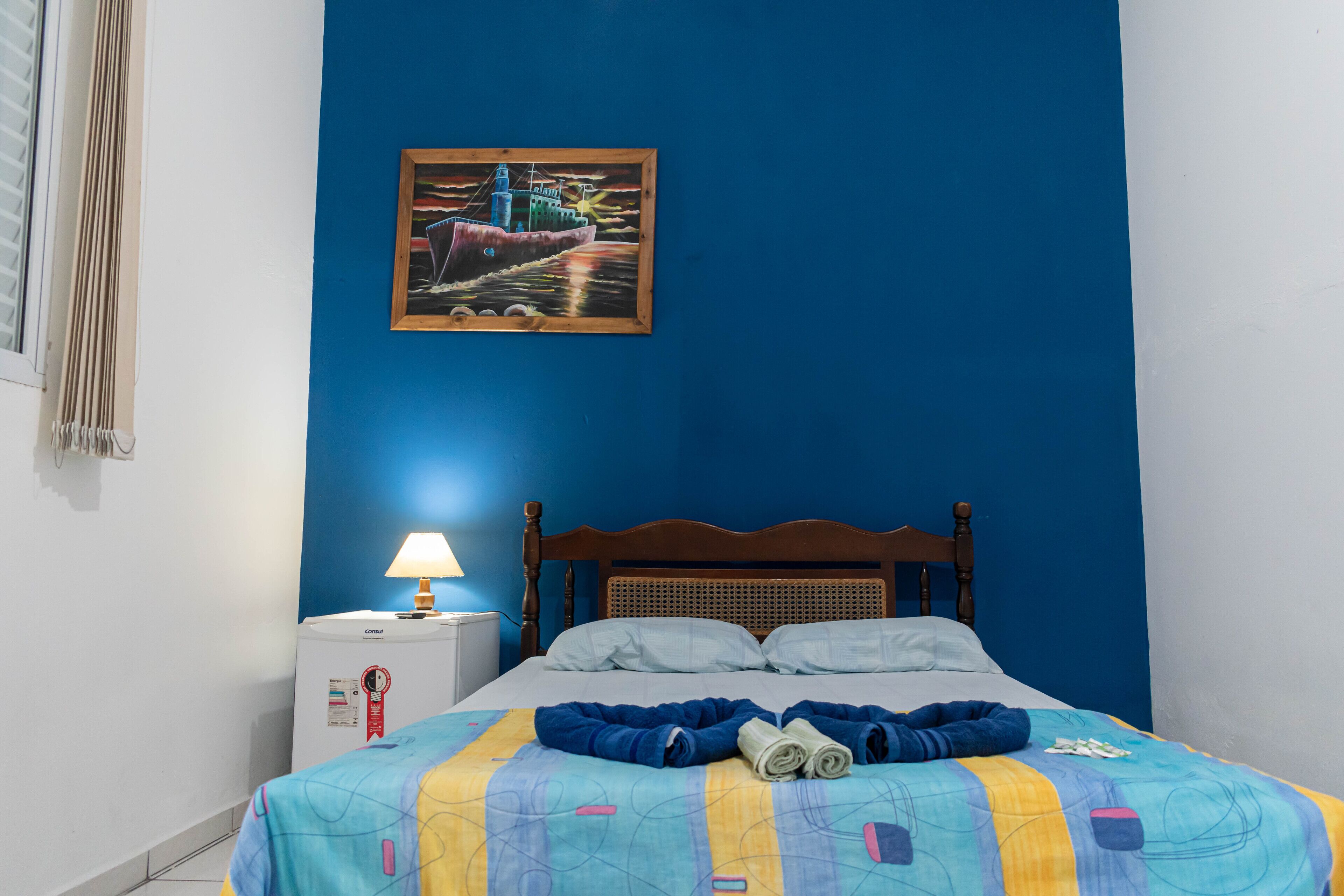 A neatly made bed with a colorful quilt, a bedside lamp, and a painting of a ship on the wall.