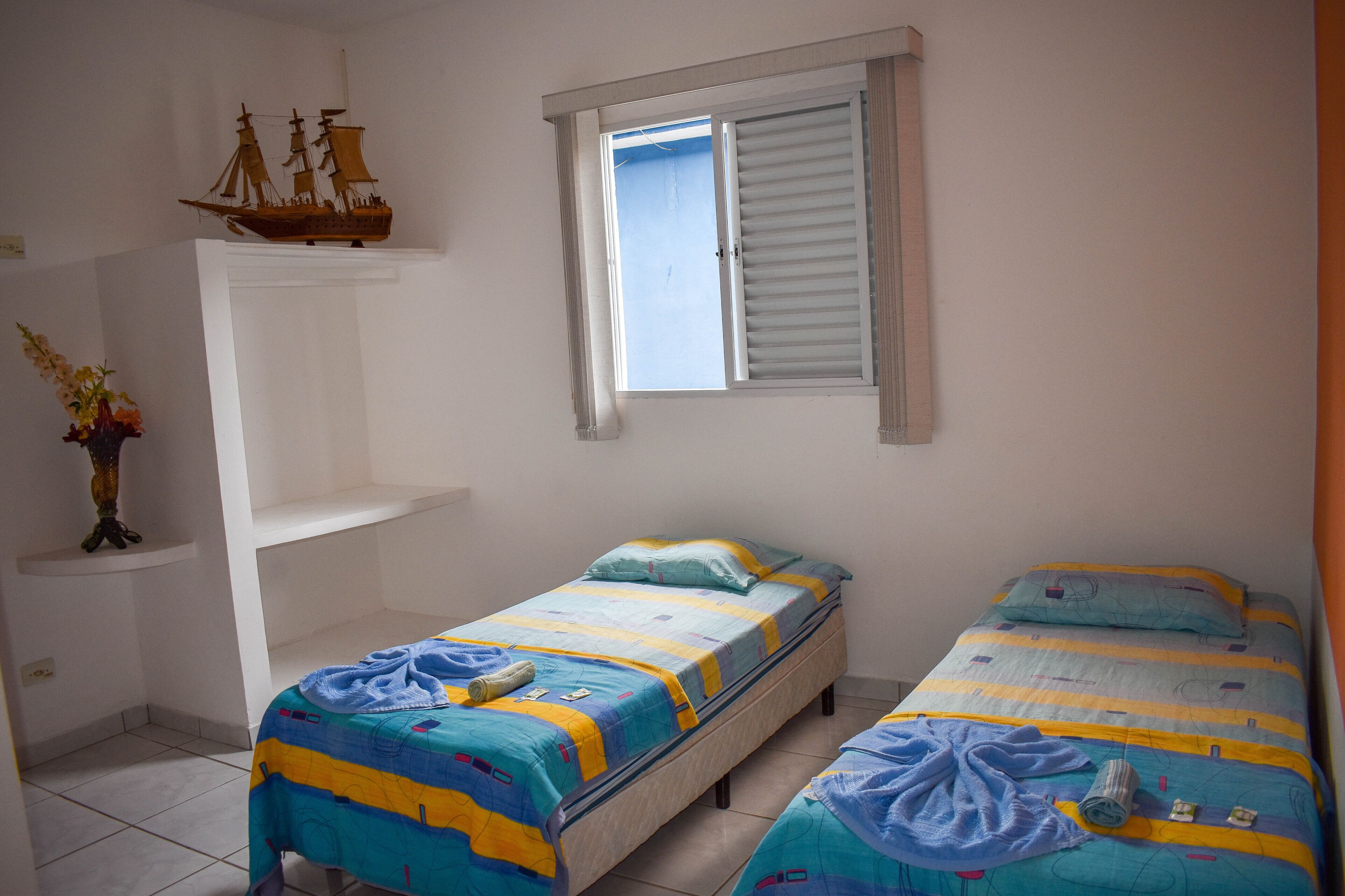 A room with two single beds, a small shelf with a ship model, and a window with shutters.