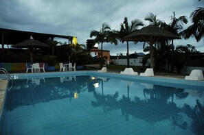 A swimming pool with tiled flooring, surrounded by white lounge chairs and umbrellas, with palm trees in the background.