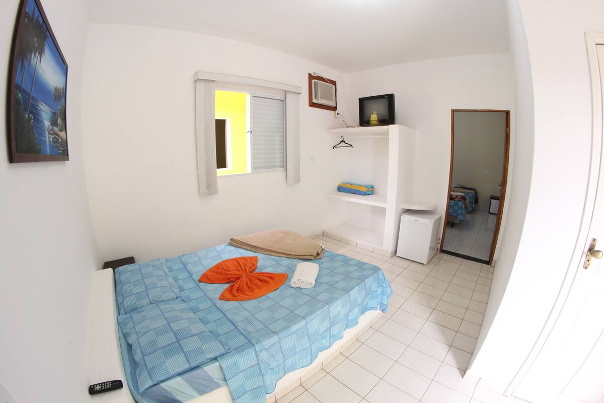 Une chambre à coucher avec un lit, une petite armoire blanche, une télévision et une fenêtre avec des stores.