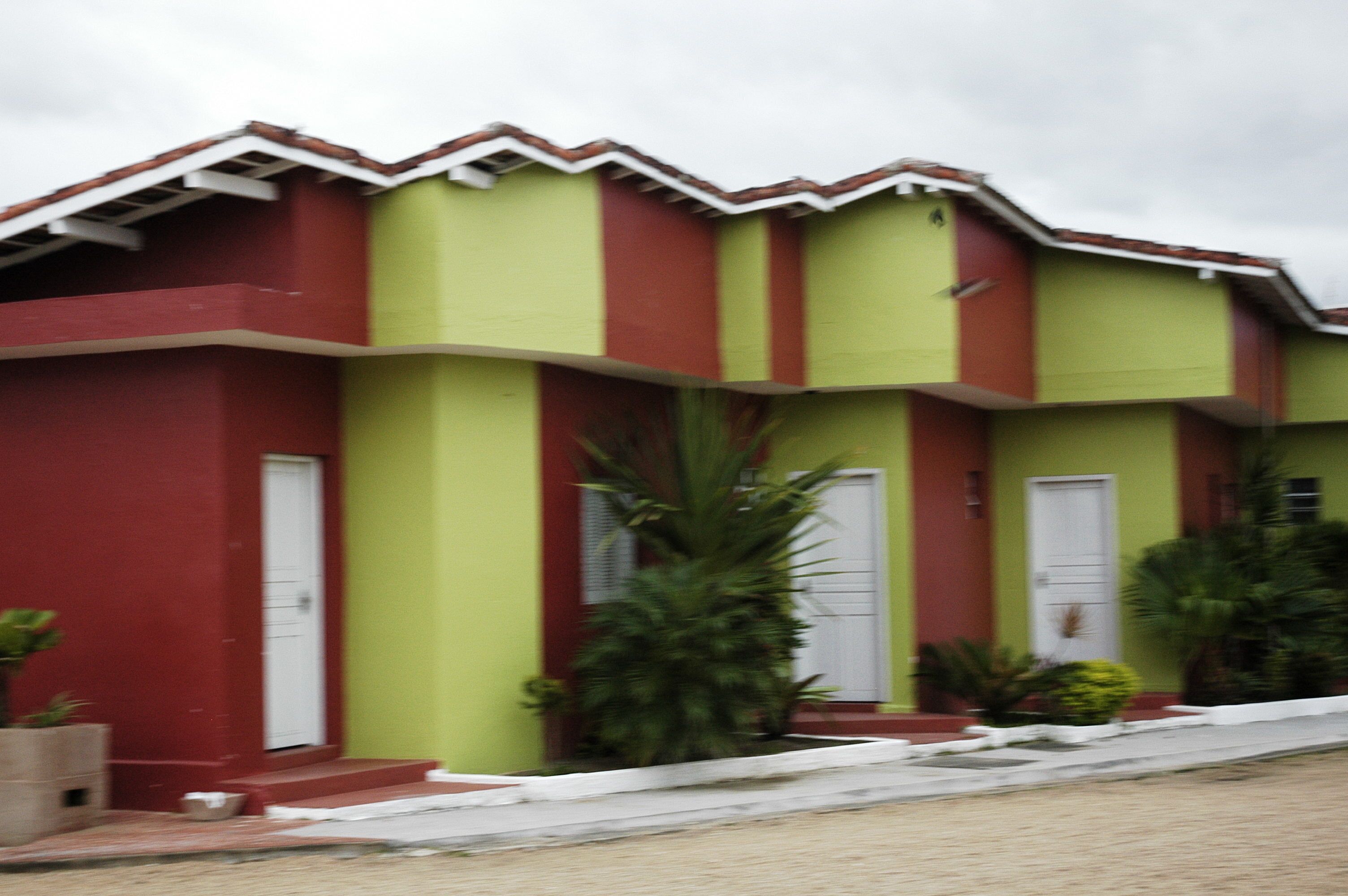 Un edificio de dos pisos con fachada roja y verde, puertas blancas y un pequeño jardín con plantas.