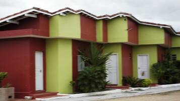Un edificio de dos pisos con fachada roja y verde, puertas blancas y un pequeño jardín con plantas.