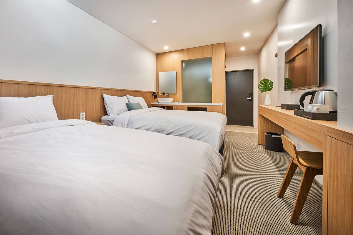 A hotel room with two beds, a desk, a mirror, a sink, and a kettle.