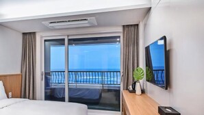 Hotelkamer met een bed, bureau, televisie en een schuifdeur naar een balkon met uitzicht op zee.