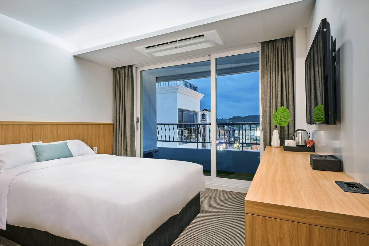 A hotel room with a bed, a wooden nightstand, a desk, and a large window with a view of a balcony and buildings.