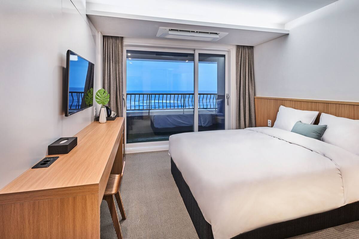A hotel room with a bed, a desk, a TV, and a balcony with a view of the sea.