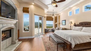A bedroom with a fireplace, a large bed, a ceiling fan, and a view of the outdoors.