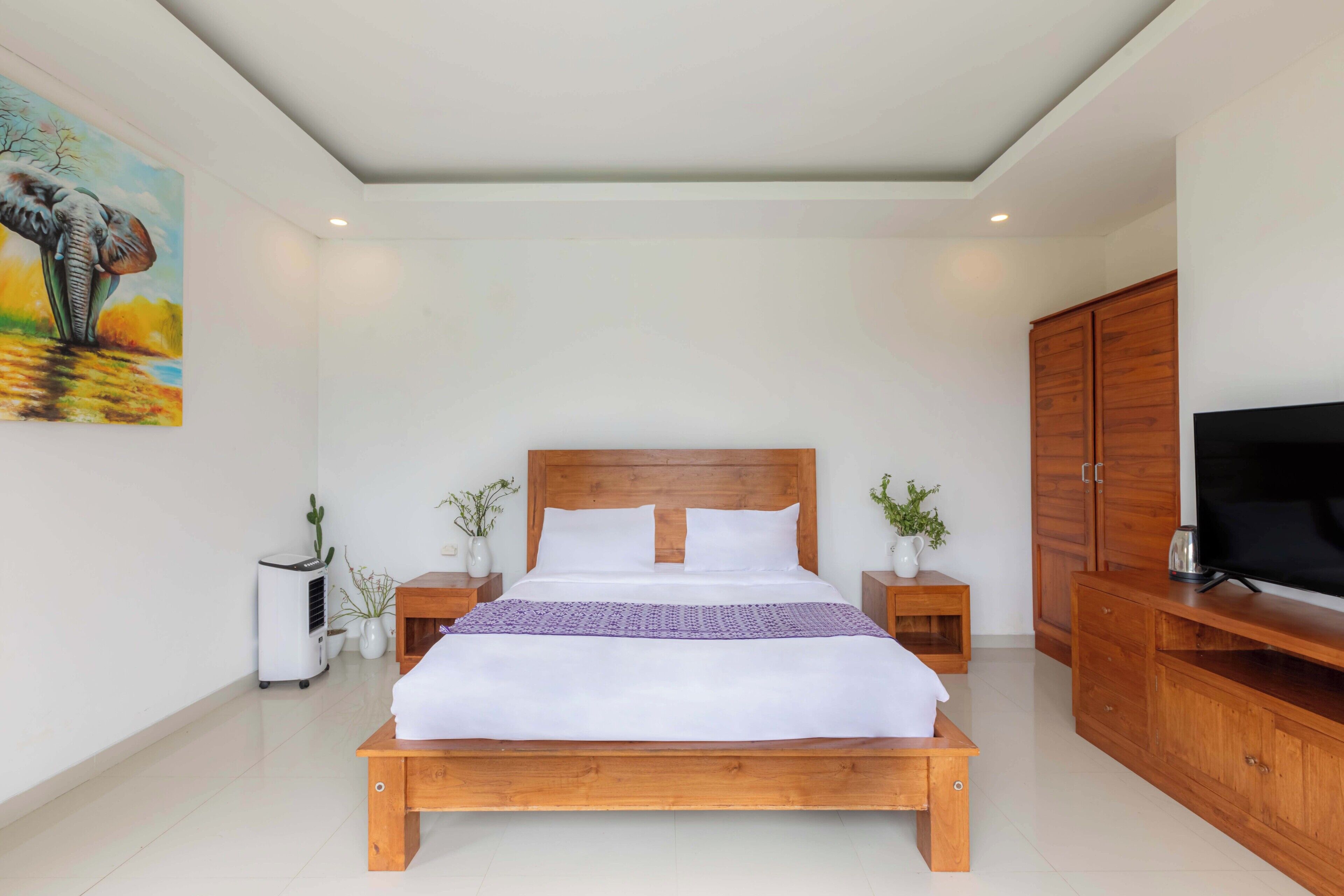 A bedroom with a wooden bed, white bedding, a television on a wooden stand, and a painting of an elephant on the wall.