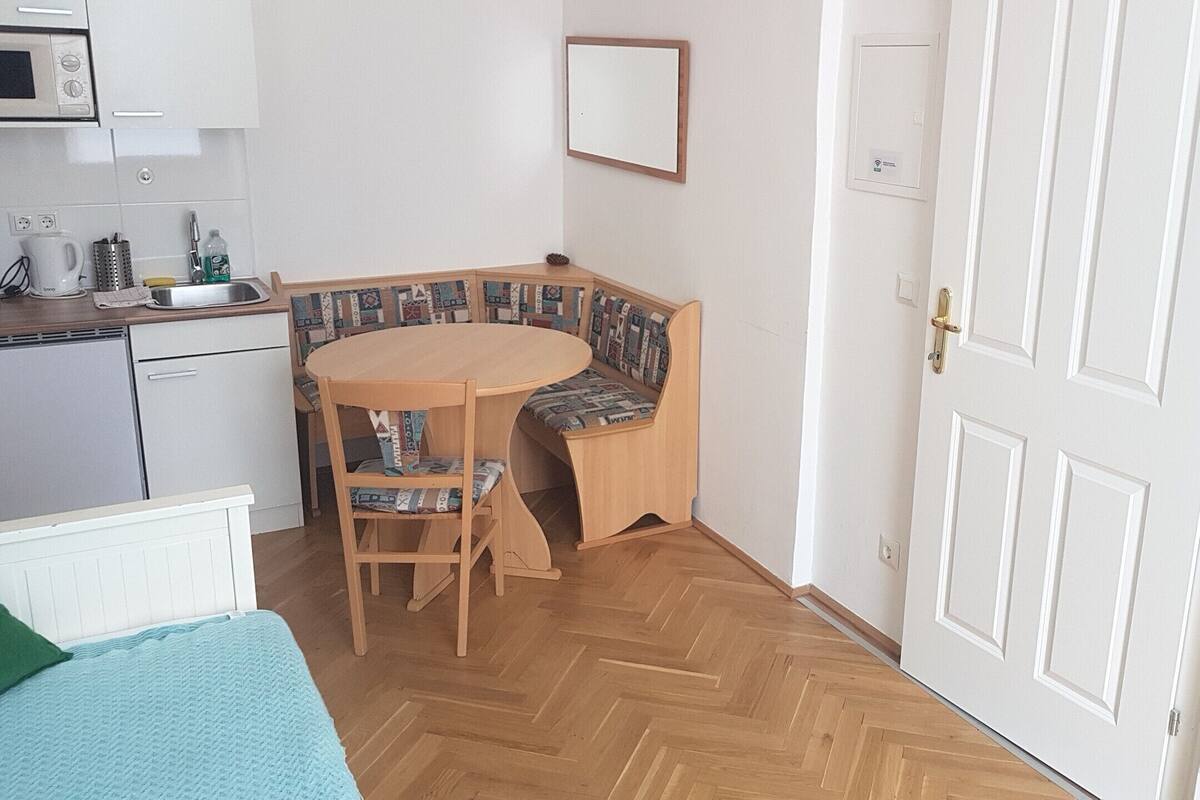 A compact kitchen with a microwave, kettle, and a sink. There's a round wooden table with a patterned cushion. The room has a herringbone wooden floor and a white door.