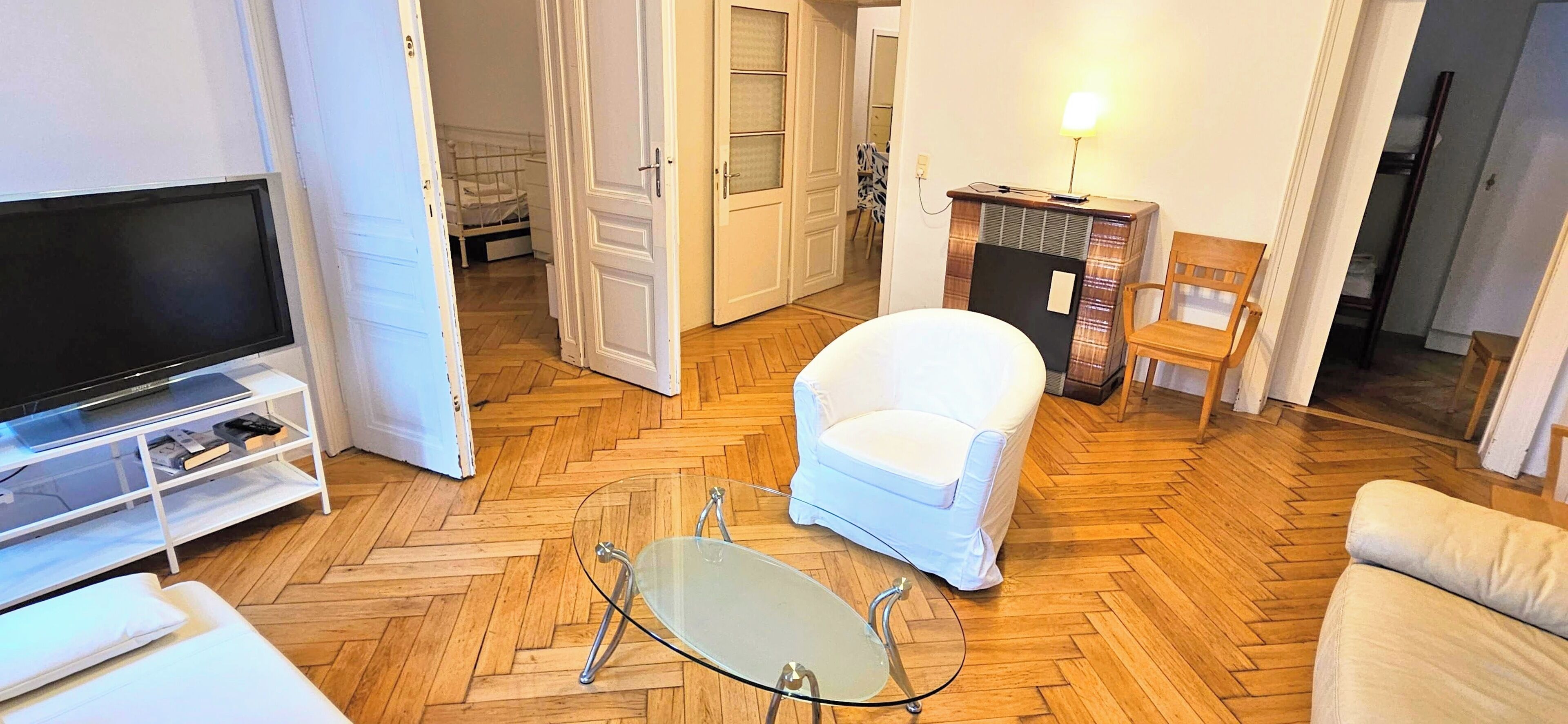 Una sala de estar con televisor, una mesa de centro de vidrio, un sillón blanco y piso de madera.